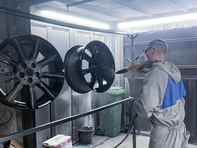 A man wearing overalls in a spray booth powder coating a set of alloy wheels.