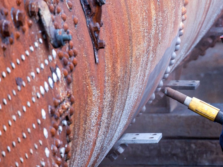 Close up of a large industrial pipe being shot blasted to remove rust.