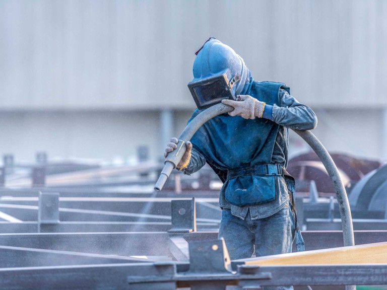 A person outside in an industrial yard shot blasting various metal items.